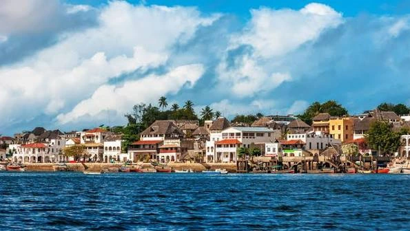 Illustration of the Lamu waterfront at sunset: cream coral-stone houses with arched windows, carved doors and a minaret, and a lateen-sailed dhow on glowing water.